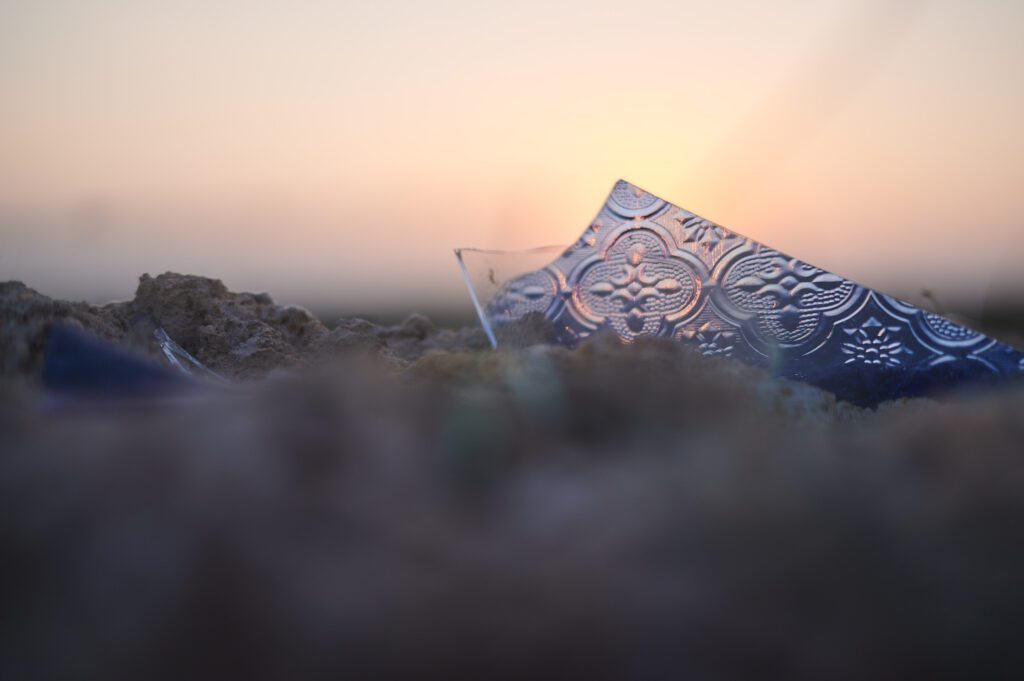 Eine Glasscherbe in Nahaufnahme, die im Sand steht und hinter der die Sonne verschwindet