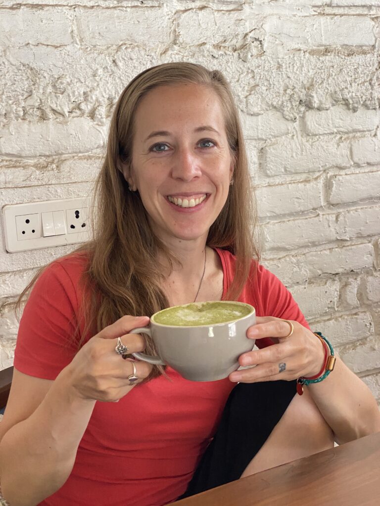 Eine Frau im roten T-Shirt lächelt in die Kamera, während sie eine Tasse mit Matcha Latte in der Hand hält. Hinter ihr ist eine Wand mit Ziegelsteinen, die weiß gestrichen sind