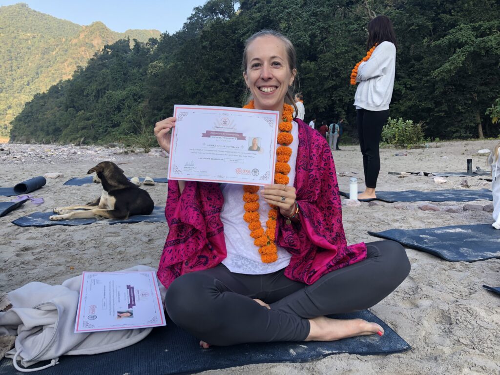 Eine blonde Frau sitzt auf einer Yogamatte, die auf Sand liegt. Sie hält ein Yoga Zertifikat in den Händen und lächelt in die Kamera. Links hinter ihr ist ein Hund zu sehen und rechts eine weitere Yoga Schülerin