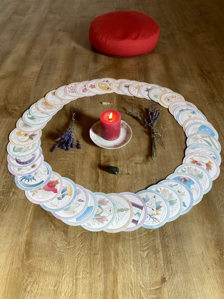 Ein Karten Deck, das im Kreis ausgerichtet ist, in dem sich eine brennende rote Kerze, Lavendel, und zwei Kristalle befinden. Hinter dem Karten Deck ist ein rotes Meditationskissen