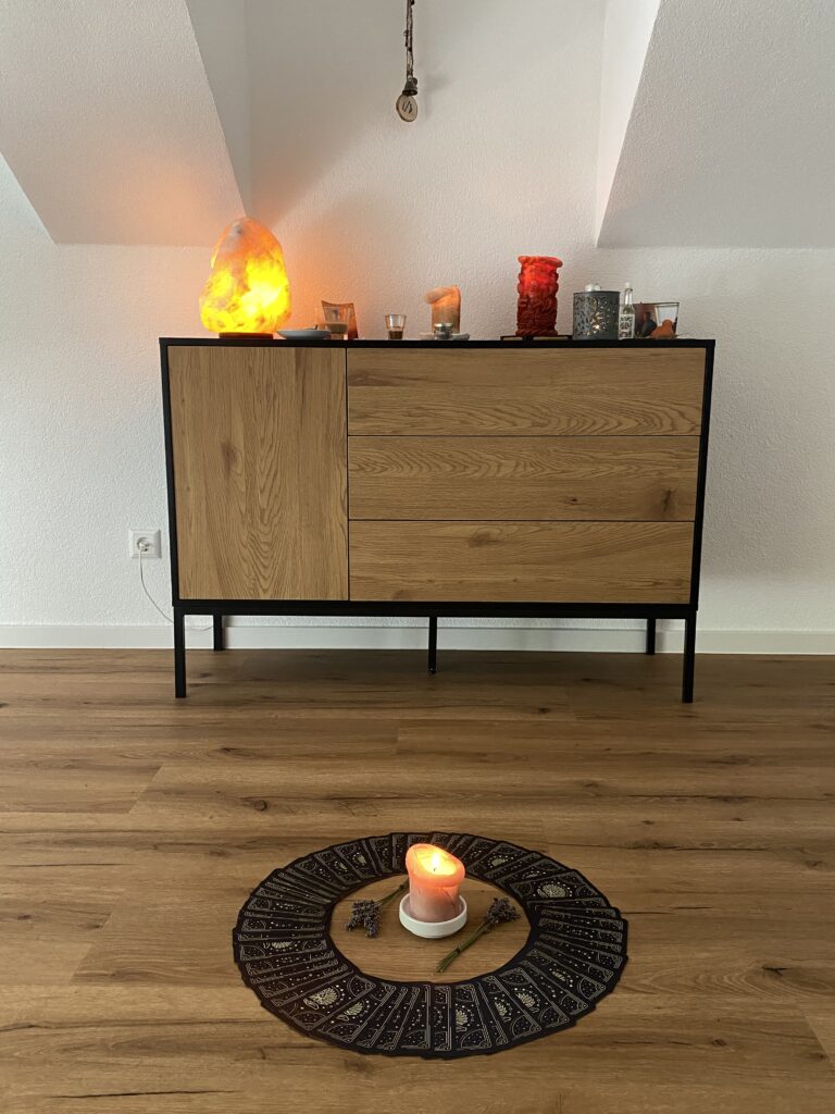 Eine Kommode auf der Kerzen und eine Steinlampe stehen. Davor ist auf dem Boden ein Kartendeck mit einer Kerze in der Mitte ausgelegt